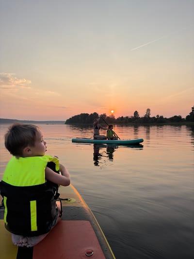 An evening scene unfolds on a wide, calm body of water, possibly a reservoir or a large river, near Ol'khovskoye, Russia. The setting sun casts a warm, orange glow across the sky, reflecting on the water's surface, creating a serene and tranquil atmosphere. A young child, facing away from the viewer and towards the water, is seated on a stand-up paddleboard, wearing a bright yellow life vest. In the distance, another stand-up paddleboard carries two individuals, one rowing and the other seated, their silhouettes visible against the sunset. The shoreline is lined with dense trees, adding depth and context to the landscape. The overall impression is one of peaceful outdoor recreation and family enjoyment.