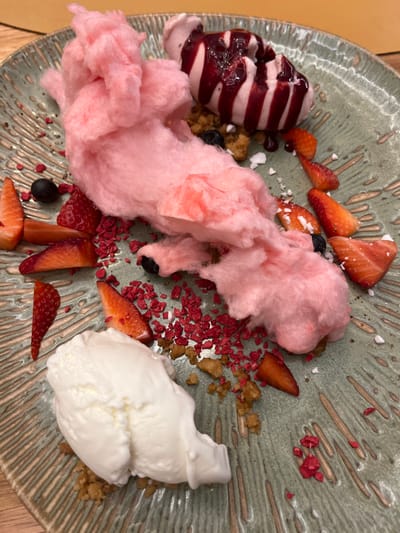 A plate of dessert is shown. It is a green, round plate with a decorative, radial design. The plate contains: a large amount of pink cotton candy; a scoop of vanilla ice cream; a scoop of pink ice cream topped with a dark red sauce; several pieces of sliced strawberry; small pieces of a brown crumb topping; a few blueberries; and several tiny, red, crispy flakes. This dessert is located at a restaurant in Las Palmas de Gran Canaria, Spain, based on the coordinates provided.