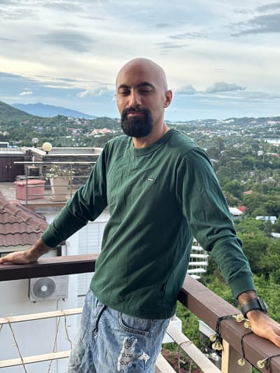 A bald, bearded man with his eyes closed is standing on a balcony overlooking a city in Ko Samui District, Thailand. He is wearing a dark green long-sleeved shirt and ripped blue jeans. The balcony has a wooden railing decorated with string lights. In the background, there are buildings, lush green trees, and mountains under a cloudy sky. An air conditioning unit is visible on the wall behind him.