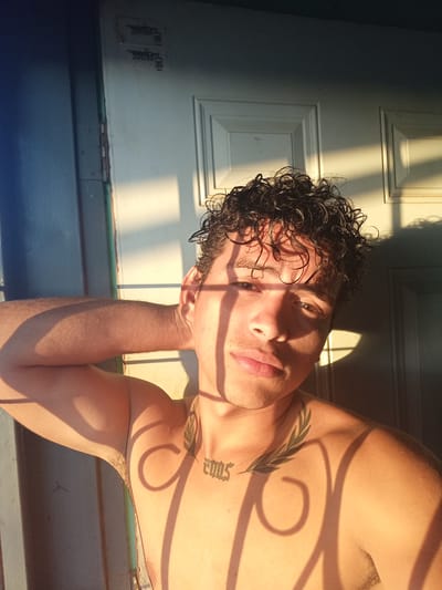 The image is a close-up, selfie-style photograph of a young man with curly dark hair and brown eyes. He is shirtless, with his left arm behind his head. He has tattoos on his chest, including a laurel wreath design on his left collarbone and the number "2005" on his chest. The lighting suggests it is either sunrise or sunset, with strong shadows cast across his face and body from what appears to be a window or a grate. He is positioned in front of a white, paneled door. The background shows a sliver of a blue wall on the left and the door on the right. The man's skin tone is light brown. There are labels on the upper part of the door that are illegible. The scene appears to be an indoor location, possibly a home, with the sunlight streaming in from an exterior source. Given the context of La Guardia, Venezuela, this location could be a residential area within the city.