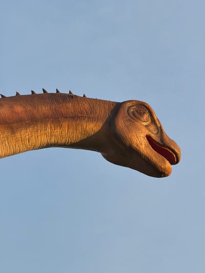 A close-up, side-view, medium shot captures the head and a portion of the neck of a large, brown, animatronic dinosaur against a clear, light blue sky. The dinosaur's textured skin is a gradient of warm browns, with highlights and shadows suggesting illumination from the right. Its mouth is open, revealing a red interior and small, visible teeth. The dinosaur's eye is detailed, with a reddish iris. Along the top of its neck, a series of pointed, dark brown scutes or spikes are visible. The background is entirely the pale blue sky, devoid of any other discernible features. The image is a professional photograph of an artificial exhibit. There is no indication of the city of Khotianivka, Ukraine in the image.