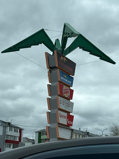 A large sign for various businesses is visible against a cloudy sky. The sign includes names such as "Oazis Plaza," "Detskiy Mir," "Burger King," "Vol shebniy Gorod," and "Krasnoe & Beloe." The sign is located in Beloyarsky, Russia. It's a horizontal image.