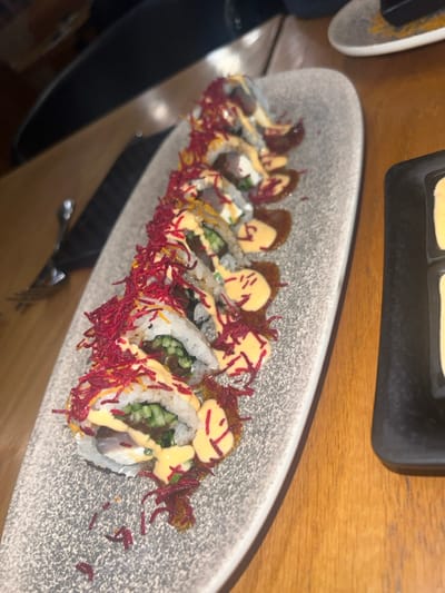 The scene depicts a long, irregularly shaped, grey-speckled ceramic plate holding eight pieces of maki-style sushi. The sushi rolls feature white rice on the exterior, with visible fillings including green cucumber strips, a darker brown protein, and a creamy white element. Each piece is generously drizzled with a light orange-yellow sauce and a darker brown sauce, then garnished with numerous thin, bright red, crispy strands, likely fried beet. The plate is positioned diagonally on a light-colored wooden table surface, extending from the bottom-left towards the top-right. In the blurred background to the left, a metal utensil and the black handle of another implement are visible, along with a dark, out-of-focus object, possibly a chair or another patron. To the right, a portion of a black, rectangular, textured dish contains two small, light yellow-beige items. The environment is an indoor dining setting, characteristic of a restaurant in Kfar Yona, Israel. No individuals are visible.