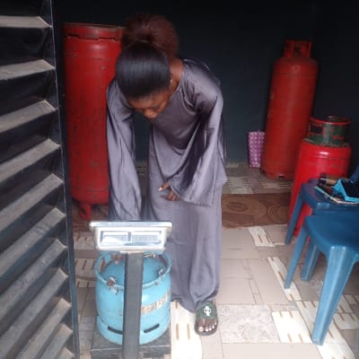 In the city of Ijotan, Nigeria, a female individual with dark hair tied back is depicted in a semi-sheltered area. She is wearing a long, grey, loose-fitting garment and dark green sandals. The individual is bending forward, focused on a silver-colored digital weighing scale with a black stand, upon which a light blue gas cylinder with visible wear is positioned. Her right hand is near the top of the cylinder. To the immediate left, a dark, vertically-slatted structure is partially visible. In the background, against a dark wall, stand two red gas cylinders of differing heights; the shorter one has a green top. A small pink polka-dotted object rests on the tiled floor between the red cylinders. To the right, a blue plastic stool holds a black smartphone and additional small objects. The floor is tiled with light-colored rectangular patterns, and a brown patterned mat is visible further back. The scene represents an individual weighing a gas cylinder at what appears to be a gas refilling or distribution point.