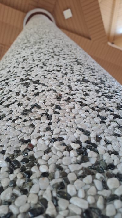 A low-angle, close-up shot captures a cylindrical column covered in a mosaic of small, smooth pebbles. The pebbles are predominantly white and black, creating a speckled texture that covers the entire surface of the column. The column appears to be made of a concrete-like material with the pebbles embedded within it. Above the column, a wooden ceiling is visible, with slats running horizontally across the frame. A single square ceiling tile is also visible near the top center of the image. The lighting suggests an indoor environment, possibly a public building or a covered walkway. The image does not depict any people or discernible actions. Without additional context, it is not possible to determine the specific location within Cotbadatunong, Indonesia.
