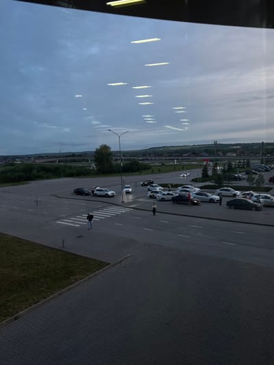 Captured from an elevated perspective, the image reveals a view of an outdoor area in Bolshoye Savino, Russia. A row of parked cars is aligned along the side of a roadway. Pedestrians are visible crossing at a designated crosswalk. In the background, structures indicative of the city are present. The sky is overcast. A section of building architecture is visible on the left side of the frame.