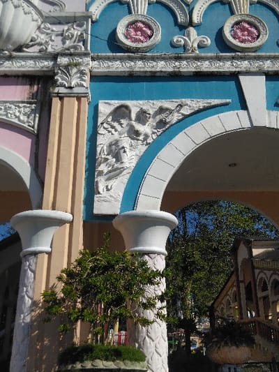 This image presents a detailed view of an ornate architectural structure in Batulawang, Indonesia. The central focus is a multi-colored facade featuring a light blue upper section and a light orange/beige lower section, adorned with extensive white decorative elements. A prominent white bas-relief sculpture of a winged, partially-draped figure holding a stringed instrument, possibly a lute, is centrally positioned above an arched opening on the right. Above this relief, the blue wall displays two circular pink rosette inserts separated by a white fleur-de-lis motif, all framed by intricate white scrollwork. To the immediate left, a fluted column, painted light orange/beige, borders a pink architectural section that includes a partial white shell-like relief on its upper portion, exhibiting some dark discoloration.
In the lower foreground, two white, fluted columns with decorative bases and flared, urn-shaped tops stand, partially obscuring a dense, potted green bush with small leaves. Through the arched opening on the right, a clear blue sky and a significant amount of green tree foliage are visible. Beyond the foliage, a section of another building or structure with multiple small, round-top arches in a reddish-brown and beige color scheme is discernible in the background. The scene is illuminated by natural light, casting distinct shadows. No individuals or specific actions are depicted within the frame.