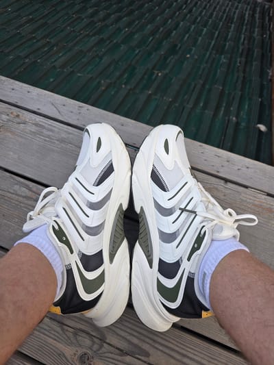 A pair of white and green athletic shoes are shown from a top-down perspective, worn by an individual sitting on a wooden dock. The shoes are positioned side-by-side with the toes pointing towards the camera. The right shoe is slightly more visible, showing a white mesh upper with layered white and dark green overlays, and a thick white sole. The left shoe mirrors the design. The person's legs, covered in white socks and displaying visible hair, are partially visible below the shoes. The background consists of weathered wooden planks, suggesting an outdoor setting, possibly near water. The environment is a wooden dock. The location is Xiaoshan District, China.