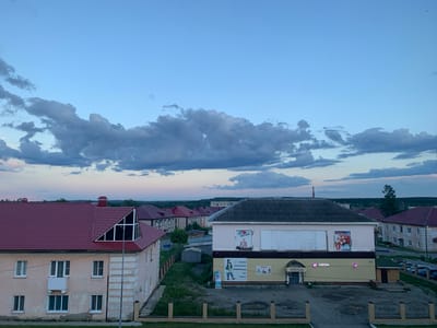 An aerial view captured at dusk showcases a residential and commercial area in Volchansk, Russia. In the foreground, a two-story building with a red tiled roof and pale yellow facade features multiple windows and a prominent triangular window on its upper floor. Adjacent to it is a larger, single-story commercial building with a dark gray roof, a beige facade, and large storefront windows displaying advertisements. A sign with "OZON" in pink illuminated letters is visible. The building also has an arched entrance labeled "РУССКИЙ ДОМ". A small kiosk with a green roof is located to the right of the commercial building. Several cars are parked in a lot behind the commercial building. The background consists of a horizon line with scattered trees and buildings, under a pale blue sky with scattered, dark gray clouds. The overall scene depicts a quiet evening in a small Russian town.