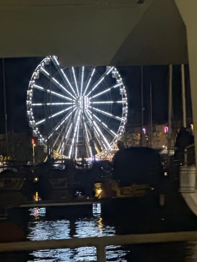 The image shows a Ferris wheel illuminated at night, likely in a city or entertainment district. The Ferris wheel is the central and most prominent object, with its spokes and carriages clearly defined by bright white lights. It is set against a dark sky, suggesting it is nighttime. In the foreground, there is water, possibly a harbor or river, with reflections of lights on its surface. Some boats or structures are visible in the water. A dark, silhouetted shape, possibly people or structures, is present in the middle ground, obscuring part of the view of the Ferris wheel and the background. The overall impression is a lively evening scene, possibly at a fair or waterfront attraction. The question of the location in the city of Agde, France, is not directly answerable from the visual information provided in the image alone.