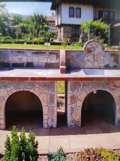 In the city of Lomets, Bulgaria, a sunlit, outdoor scene features a meticulously constructed stone barbeque and seating area. The barbeque is an impressive structure, divided into two equal sections, each with a prominent arched opening below. The entire structure is adorned with a mosaic of irregular, flat stones in varying shades of tan, gray, and dark brown, outlined with dark grout, creating a visually striking pattern. Red brick is visible as an accent, particularly along the top edges of the barbeque and as a border.

Above the barbeque, integrated stone benches with rounded, decorative backs flank a central divide. The left bench has a simple flat back, while the right bench features a curved, almost semi-circular stone element. The surface of the barbeque appears to be a grill grate.

The barbeque sits on a patio paved with large, light-colored rectangular stones, bordered by a thin red brick edge. In front of the barbeque, low-lying green shrubs and other ground cover plants are neatly arranged, adding a touch of natural beauty to the manicured outdoor space.

Behind the barbeque, traditional Bulgarian architecture is visible. A white-washed house with a red-tiled roof and dark-framed windows stands prominently. The house exhibits the characteristic construction style of the region, with stone foundations and upper levels. Lush green trees and vegetation surround the house, indicating a well-maintained yard and garden. The sky is a clear, bright blue, suggesting a warm, sunny day. The overall impression is one of a charming, well-appointed outdoor living space, likely designed for cooking and socializing.