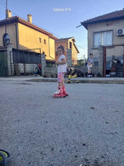 Two young girls are on a residential street in Obrochishte, Bulgaria. One girl is wearing pink roller skates, a white t-shirt, and patterned shorts. She is standing in the street and facing the camera. The other girl is slightly further away, near a building, holding a round yellow object, and also appears to be wearing roller skates. The street is asphalt, and the background includes houses, a yellow scooter, and some stacked wood. The weather appears sunny and clear. The image is taken from a mobile phone, with the "Galaxy A15" watermark present on the left side.