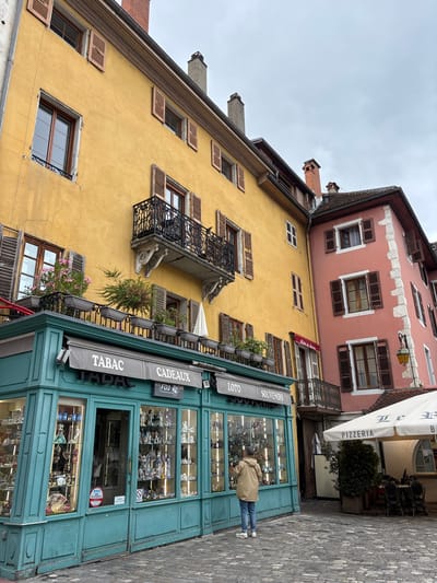 In Annecy, France, a person stands before a teal-colored storefront displaying an assortment of figurines and souvenirs. The storefront is adorned with awnings labeled "TABAC," "CADEAUX," "LOTO," and "SOUVENIRS." Adjacent to this shop, a pink building features windows with brown shutters, and beyond it, a yellow building showcases similar architectural details. The scene unfolds on a cobblestone street, with an outdoor seating area for a "PIZZERIA" visible to the right, covered by a white umbrella. The sky is overcast.