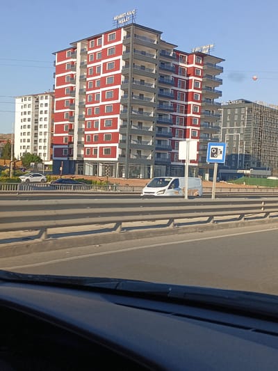 The image depicts a modern apartment complex with red and white facades, located in Yahşihan, Turkey. A white van with orange lettering is parked on the road in front of the complex. A traffic sign indicating a camera is also visible. The scene is captured from a moving vehicle, as suggested by the blurred foreground. The background shows other buildings, a clear sky, and power lines.