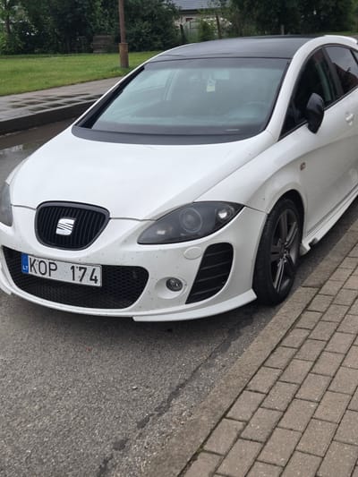 A white SEAT Leon hatchback is parked on a brick-paved sidewalk next to a grassy area. The car is facing slightly towards the right of the frame, with its front bumper and driver's side prominently visible. The car has a black grille with the SEAT logo in the center, and a license plate reading "KOP 174 LT" is attached to the front. The headlights are dark, and the front wheel is a dark alloy. The car appears to be a bit dusty, with specks of dirt on its white paint.

The background shows a grassy area with some trees and a hint of a building on the left. The sidewalk is made of rectangular bricks. The scene appears to be outdoors, possibly in a residential or public area. The lighting suggests it might be daytime, and the presence of water on the ground and a slightly overcast sky could indicate recent rain or damp conditions. There are no people visible in the image. The overall impression is of a parked car in an urban or suburban setting in Širvintos, Lithuania.