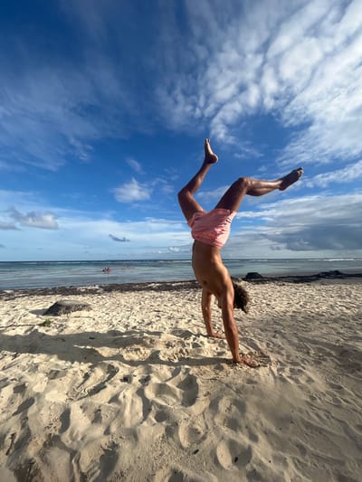 A man in pink shorts is positioned horizontally in front of a beach and blue sky with scattered clouds. His right hand touches the sand on the beach, while his legs extend upwards, with his feet pointing towards the sky. The beach stretches from the bottom right to the middle of the image. Beyond the beach, there's a strip of ocean that runs horizontally through the center of the image. The sky with clouds fills the upper left area. The photo appears to have been rotated 90 degrees counter-clockwise. It's difficult to determine the exact location in Sainte-Anne, France without more contextual information.