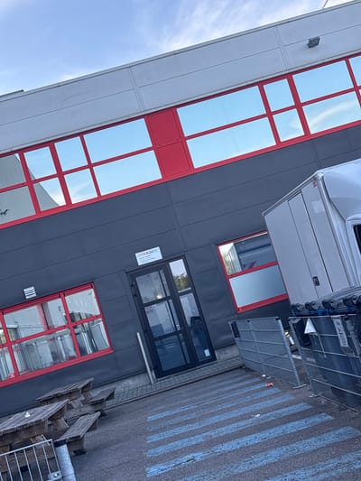 The image depicts the exterior of a modern industrial building with a dark gray facade and bright red window frames. The building has large, reflective windows that mirror the blue sky and some clouds. Above the dark gray section, there is a lighter gray upper level with a horizontal band. There is a glass entrance door on the dark gray section, with a small sign above it that reads "DYNALOGIC". A white truck with a cargo box is parked to the right of the entrance, partially obscuring some windows. In front of the building, there are wooden picnic tables and benches. The ground in front of the building is asphalt with blue painted stripes, possibly indicating a pedestrian crossing or parking area. Metal fencing is visible in the foreground and next to the truck. A single red and white can is on the ground near the fence. The scene appears to be in an industrial or commercial area, with a clear sky suggesting daytime. The image does not show any people. The specific location in the city of Boom, Belgium, is not directly indicated by any visible elements within the image itself.