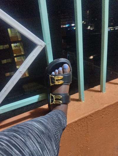 A person's leg, clad in gray striped pants, rests on a ledge next to a railing. The foot wears a black sandal with two wide straps, each adorned with a gold-colored buckle. Toes are painted a light purple. A railing of green vertical bars and white angled bars is visible in the background. Outside the railing, the blurry lights of a city at night shine in the distance. The ledge appears to be a tan or orange color.