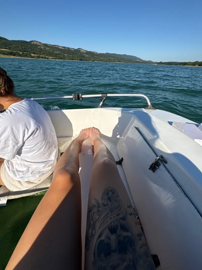 The image shows two individuals on a boat traveling on a body of water. One individual, seen from the back, wears a white t-shirt and light-colored shorts. The other individual's legs are prominently featured in the foreground, with bare feet and a tattoo visible on the left thigh. The boat appears to be moving, as evidenced by the wake in the water. The background consists of a hilly, tree-covered landscape under a clear blue sky. The location is identified as Golyama Zhelyazna, Bulgaria.