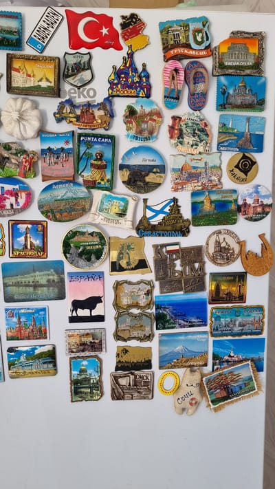 The image displays a collection of refrigerator magnets, predominantly featuring architectural landmarks and cultural symbols from various global locations. The magnets are arranged on a white, flat surface, likely the door of a refrigerator. Visible magnets represent cities and regions such as Thailand, Aspen, Moscow, Cyprus, Yerevan Opera, Sevastopol, Armenia, Florence, Spain, Dubai, Samara, and Santo Domingo. One magnet depicts a bull and the word "ESPAÑA," suggesting a connection to Spain. Another magnet shows a stylized image of the city of Moscow with its iconic red domes. A magnet with a Turkish flag is also present. The overall arrangement suggests a personal collection of travel souvenirs. There are no people, actions, or interactions depicted in the image. The environment is a plain white surface. There is no visible information or imagery within the collection of magnets that specifically relates to the city of Chereshnya, Russia.