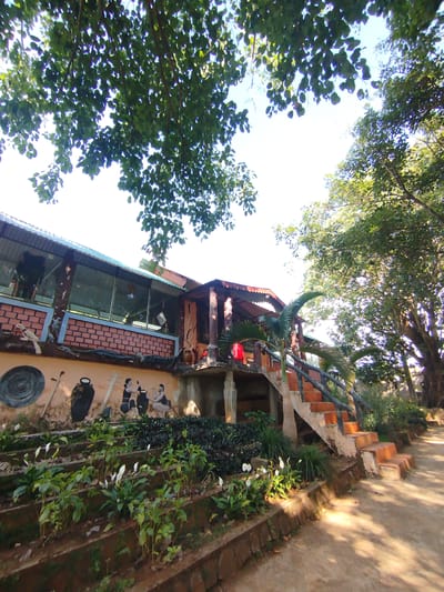 The image shows a building with a brick and stucco exterior, located in An Phu Ward, Vietnam. A covered patio area is on the left, enclosed with large windows. The stucco section features a mural depicting people and objects, including pots and musical instruments. A set of brick stairs with a log railing leads up to the patio. In front of the building are terraced gardens with flowering plants. Trees overhang the building, casting shadows. The ground is dirt and sunlight is visible.