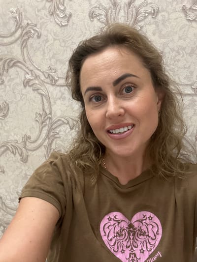 A woman is pictured from the chest up, smiling into the camera. She has light brown hair styled in loose curls, blue eyes, and fair skin. She is wearing a brown t-shirt with a pink heart design on the front, and a thin gold chain necklace. The background is a wall with a light-colored, ornate wallpaper pattern. The image was taken indoors.