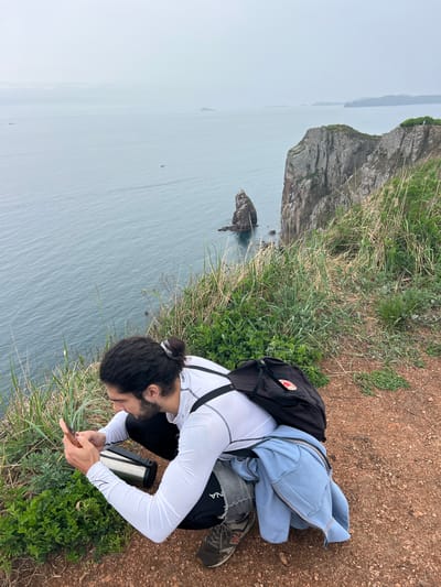 A man is crouched on a grassy, earthen cliff edge in Russkiy Island, Russia, overlooking the sea. He is focused on his mobile phone, his dark hair pulled back in a bun. He wears a white long-sleeved shirt, dark pants, and a black backpack. A light blue jacket lies near him. In the background, a rocky cliff juts out into the water. The sea is grey and misty. A metal container sits beside him.