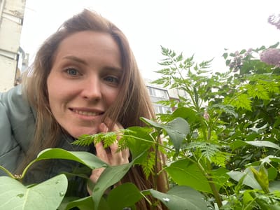 A medium shot shows a woman with long brown hair and blue eyes, looking directly at the camera with a slight smile. She is wearing a light green puffer jacket. Her right hand is touching her face near her mouth, with her fingers partially obscuring a feathery green leaf. The background consists of lush green foliage, including some larger leaves and smaller, fern-like leaves, and hints of pink blossoms in the upper right corner. In the distance, there are buildings, suggesting an urban environment. The lighting is overcast, indicating a cloudy day. The scene appears to be outdoors, possibly in a park or garden area in Yuzhno-Sakhalinsk, Russia, given the architectural style of the distant buildings which resembles those found in Russian cities.