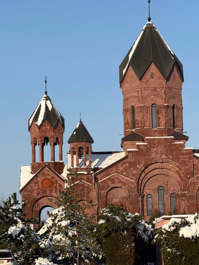 The image depicts the Surp Hovhannes Mkrtich (Saint John the Baptist) Armenian Apostolic Church in Rassvet, Russia, under a clear, bright blue sky during winter. The prominent structure is constructed from reddish-brown brick or stone, displaying intricate architectural details.
The church features a large central conical dome with a dark, segmented roof, topped by a slender cross finial. This main dome section includes tall, narrow arched windows. To the left of the central dome, two smaller, open bell towers with dark conical roofs are visible, each also topped with a cross finial. These bell towers feature arched openings supported by columns. An additional smaller, closed dome is partially visible behind these bell towers. All visible roofs and various architectural ledges on the building are partially covered with white snow. Decorative carvings, including cruciform designs, are discernible on the church's facade and dome bases.
In the foreground, a dense collection of evergreen trees and bushes, heavily covered with white snow, partially obscures the lower sections of the church. A portion of a white vehicle, possibly an SUV, with a partial license plate ending in "...782," is visible in the bottom-left corner, largely obscured by the snow-laden foliage. The scene is illuminated by strong, directional sunlight, indicating a bright winter day. No individuals or active interactions are present in the frame.