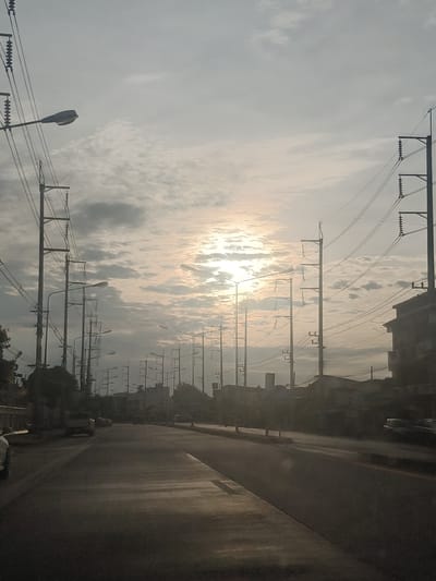 A low-angle, vertical shot captures a hazy, sunlit sky over a street lined with utility poles and overhead wires. The sun is partially obscured by clouds, creating a diffused light across the scene. Multiple streetlights, some visible on poles, are present along the road. Several cars are seen on the road, with buildings and structures lining the right side of the street. The road itself is paved, with visible lane markings. The overall impression is that of a street scene in a tropical or subtropical city during daylight hours, with atmospheric conditions affecting visibility. The image is consistent with a view captured within Chiang Rai City Municipality, Thailand, given the common urban infrastructure and lighting seen.