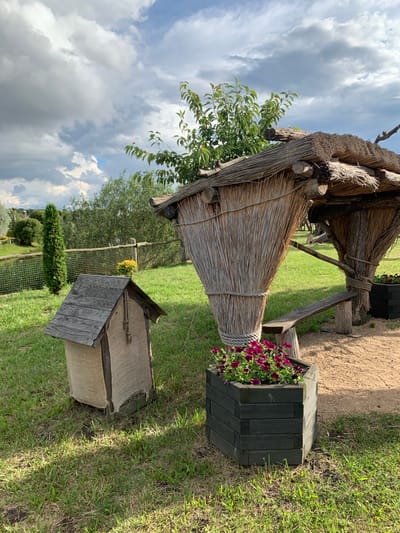 The image depicts an outdoor scene in the Малахавецкі сельскі Савет, Belarus, featuring rustic structures and greenery. In the foreground, a small, wooden, house-like object with a peaked roof and canvas sides sits on a grassy area. To its right, a hexagonal wooden planter filled with vibrant purple flowers is placed on sandy ground. Behind these objects, a larger, thatched structure with a cone-shaped body is supported by tree trunks and a wooden bench. This structure is tied with rope. In the background, a pond is visible beyond a wire fence, with a tall, slender evergreen tree standing near its edge. Lush green grass covers most of the ground, and trees and bushes form the backdrop under a cloudy sky with patches of blue.