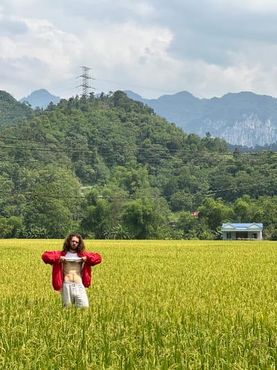 The image, taken in Ngọc Đường Commune, Vietnam, shows a man posing in a vibrant rice paddy field against a backdrop of lush mountains and an overcast sky. The man, with long, dark curly hair and a beard, stands in the foreground, wearing a red open jacket, a white t-shirt, and light-colored pants. He is actively lifting his white t-shirt to reveal his abdomen, looking directly at the camera with a neutral expression. Some faint, indiscernible text is visible on his t-shirt.
The extensive rice paddy field, filled with green and yellow rice plants, occupies the lower portion of the frame, stretching towards the distant mountains. In the mid-ground, a large electricity pylon with multiple power lines traverses the scene, positioned against the green, tree-covered slopes. Further into the background, rolling mountains are covered with dense foliage, appearing hazier due to the distance. A small, light-colored house with a distinctive blue roof is nestled among the trees on the right side of the mountain range. The sky above is overcast with soft, grey clouds, indicating daytime with diffused light. The overall scene blends natural agricultural landscapes with elements of human infrastructure in a rural setting.