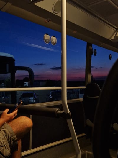 The image depicts the interior of a bus at dusk, with a view of the twilight sky through the window. The sky exhibits a gradient of deep blue to shades of pink and purple. Outside, a landscape of fields and a road with several vehicles, including cars and a truck, are visible. The interior of the bus shows a hand holding a smartphone, with part of a person's leg visible. A white pole, likely a support for passengers, runs vertically through the frame. Two circular stickers are affixed to the window; one depicts a stroller, and the other a person wearing a seatbelt. The specific location in the city of Bahovitsa, Bulgaria, is not discernable from the image itself.