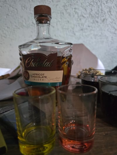 The image shows a bottle of "Creme de la Creme Chocolat - APRICOT CHOCOLATE - BLEND" next to two glasses. One glass appears to contain a reddish liquid, while the other contains a yellow liquid. The bottle is made of clear glass with a wooden cork stopper. The label on the bottle features elegant script and a circular emblem. The scene is set on a wooden surface, possibly a table, with a textured white surface in the background. The lighting suggests an indoor setting, and the time of day is not clearly discernible. No people are visible in the image. The location context is Yevpatoriya, Ukraine.