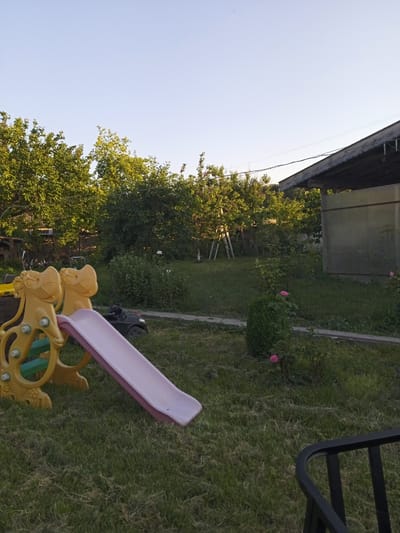 The image depicts a backyard scene in Vehtovo, Bulgaria. In the foreground, a yellow plastic slide with a pink ramp is situated on a patch of grass. To the left of the slide, a portion of a yellow plastic structure resembling a toy animal is visible. Behind the slide, a small black toy car is parked on the grass. The background features lush green trees and bushes, with a light-colored building with a corrugated roof on the right side of the frame. A metal ladder is leaning against one of the trees. The sky is clear and pale blue. The overall impression is of a quiet, residential outdoor space.