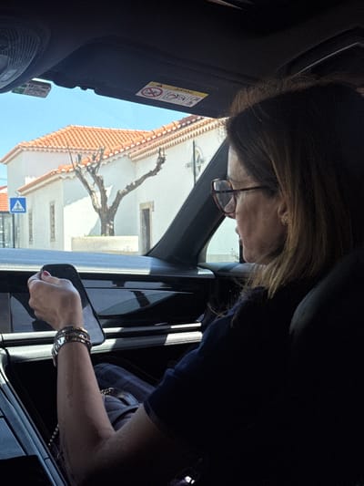 A high-angle shot captures a person, likely a woman, leaning out of a vehicle's open window. She has brown hair, wears glasses, and an earring is visible on her right ear. Her right arm, adorned with multiple bracelets and a watch, extends out of the window, holding a smartphone in her hand. She appears to be looking down and interacting with the phone or something outside the vehicle.
The interior of the vehicle shows parts of a dark fabric seat, the door frame, and a sun visor with an "AIRBAG" warning sticker and related symbols.
Through the open window, a sunny outdoor scene is visible under a clear blue sky, suggesting it is midday or early afternoon. A building with a traditional orange tiled roof and white walls occupies the background. A large, bare tree branch extends across the view in front of the building. Near the bottom left, a blue triangular road sign with a pedestrian crossing symbol is partially visible. The bright light casts distinct shadows. The setting appears to be a street in an urban or semi-urban area, consistent with Torres Vedras, Portugal.