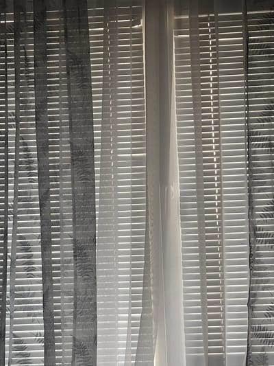The image captures a close-up, static view of a modern window covering, likely a 'day and night' or 'zebra' blind, from an interior perspective. The subject is the intricate detail and light-filtering effect of this window treatment.
The covering is composed of alternating horizontal bands. Some bands consist of translucent white material, appearing as a series of narrow, vertically oriented slats or pleats. Through these white sections, bright, direct daylight intensely penetrates, creating a distinct pattern of vertical lines of light and shadow. Interspersed with these bright white bands are wider horizontal strips of sheer fabric. These fabric strips are patterned with a repeating botanical motif, resembling fern fronds or similar foliage, rendered in shades of grey against a lighter grey background.
The strong backlighting indicates a bright, sunny daytime, with sunlight directly filtering through the window. This creates a high-contrast visual effect, highlighting the texture and design of the blinds. There is no discernible activity or event taking place; the image is a still life focused on the interplay of light, shadow, and material.
Notable details include the crisp, clean lines of the vertical slats and the softer, more diffused light coming through the patterned fabric. The overall color palette is limited to whites, greys, and shadows. No people or specific objects other than the window covering are visible, and there is no visible text. The image provides no specific visual cues to its location, although it is contextually stated to be Novi Pazar, Bulgaria.