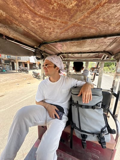 A man wearing a white outfit and bandana sits in the back of a three-wheeled auto-rickshaw taxi. He looks out the left side of the vehicle, holding a cell phone. A grey backpack is next to him. The driver of the auto-rickshaw is visible in the rearview mirror. The taxi appears to be traveling on a street in Mathura, India, based on the architecture and business signage visible outside. Several motorcycles are parked nearby.