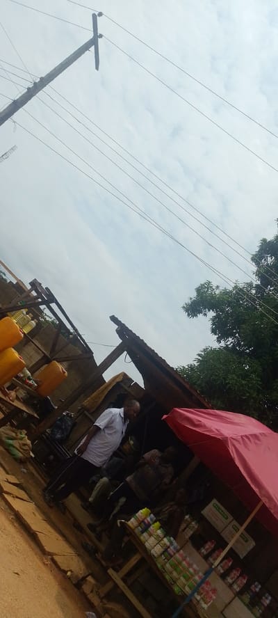 A tilted, medium shot captures an outdoor market scene in Baissa, Nigeria. In the foreground, a dirt path with visible cracks runs diagonally across the frame. To the right, a wooden stall displays rows of bottled products, some with labels in green and white packaging. Behind the stall, a man in a patterned shirt sits partially obscured by a large pink umbrella. To the left of the stall, a man in a white short-sleeved shirt and dark pants stands in profile, looking down and to his left. Behind him, stacked yellow jerrycans and several bottles of liquid are visible on wooden shelves. A partially constructed wooden structure with a corrugated roof covers part of the scene. Overhead, multiple power lines crisscross against a cloudy sky, with a wooden utility pole visible in the upper left quadrant. Lush green trees frame the right side of the image.