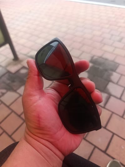 A person's hand is holding a pair of black-framed sunglasses with dark lenses. The sunglasses are partially opened, revealing a reddish tint inside the lenses. The hand appears to be holding the sunglasses by the frame. The background consists of a tiled surface, likely a walkway or patio, with light brown and grey square tiles. A dark vertical object, possibly a pole or support, is visible to the left of the hand. The scene is outdoors, and the lighting suggests it is daytime. There are no specific landmarks or environmental cues to definitively place this image in Fanipal, Belarus.