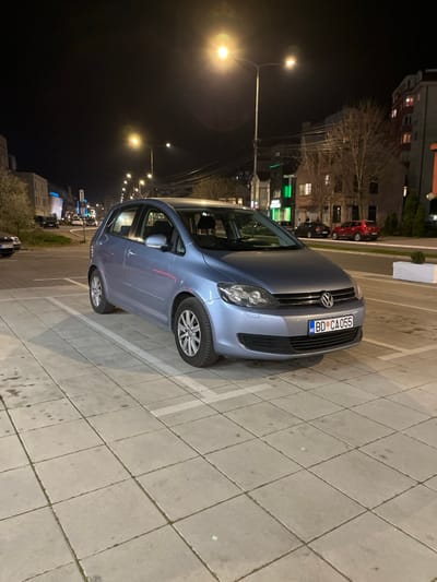 The image depicts a nighttime urban street scene in Niš, Serbia, specifically on Bulevar Nemanjića. A light blue Volkswagen Golf Plus hatchback is the primary subject, parked facing slightly right towards the viewer in a designated parking space. The car bears a Montenegrin license plate, "BD CA055," indicating its registration in Budva, Montenegro. The vehicle appears to be in clean condition.
The parking area consists of light grey square paving slabs, marked with white lines defining the parking space. Some stains are visible on the pavement.
In the background, a multi-lane asphalt road is visible, illuminated by multiple streetlights emitting a warm yellow-orange light. On the left side of the road, several multi-story buildings line the street, with one distant building displaying blue illumination. Another dark-colored vehicle is partially visible parked on the far left. On the right side of the road, commercial and residential buildings are present. A prominent building features a brightly illuminated green sign on its ground floor. Further down, a multi-story residential building has several illuminated windows. Deciduous trees with bare branches, suggesting late autumn or winter, stand between the buildings. A white rectangular planter box containing a small plant separates the parking area from the roadside on the right. Two additional vehicles, a dark sedan and a red SUV, are parked further down the street on the right. No people are visible in the scene, and no active interactions or movements are depicted.