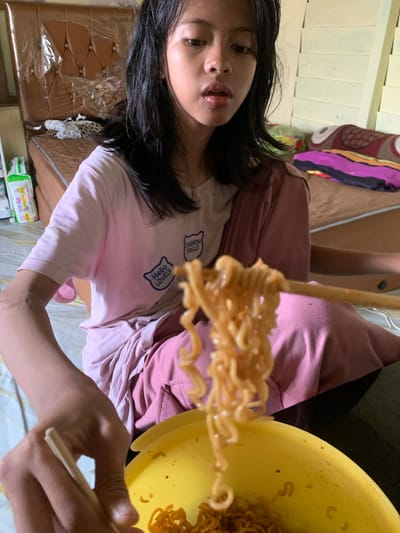 A young female with dark hair, wearing a pink t-shirt with graphics, is sitting on a bed and eating instant noodles with chopsticks. She is in a bedroom in Sungai Meriam, Indonesia. The bed has a brown headboard covered in plastic wrap, and there are pillows and a blanket on the bed. A yellow bowl of noodles is visible in the foreground.