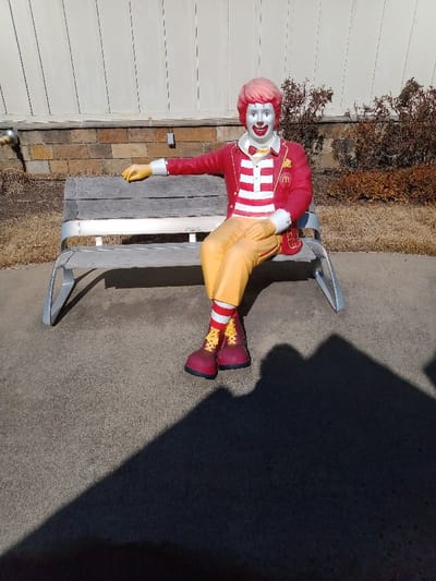 A statue of Ronald McDonald sits on a gray bench outside. The statue wears a red and white striped shirt, yellow pants, red jacket, red and white socks, and red shoes. A brown and yellow brick wall, along with white siding is in the background. A large shadow is present on the concrete floor. A row of brown shrubs is also visible.