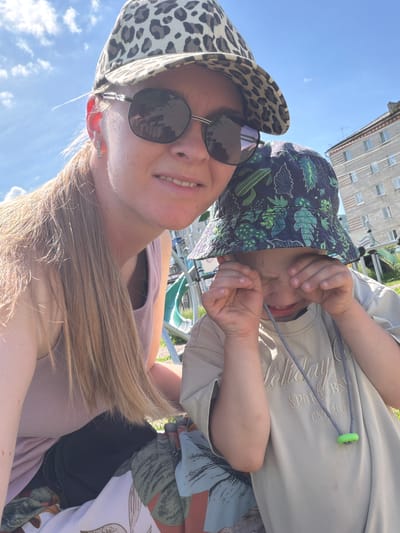 A woman and a child are taking a selfie outdoors on a sunny day, with a residential building and playground equipment visible in the background. The woman, on the left, is wearing a leopard-print baseball cap, sunglasses, and a pink tank top. Her blonde hair is long and loose. The child, on the right, is wearing a bucket hat with a cactus print and a light-colored t-shirt. The child is rubbing their eyes. They appear to be sitting on a blanket with a floral pattern. The location is identified as being in the city of Kuldur, Russia.