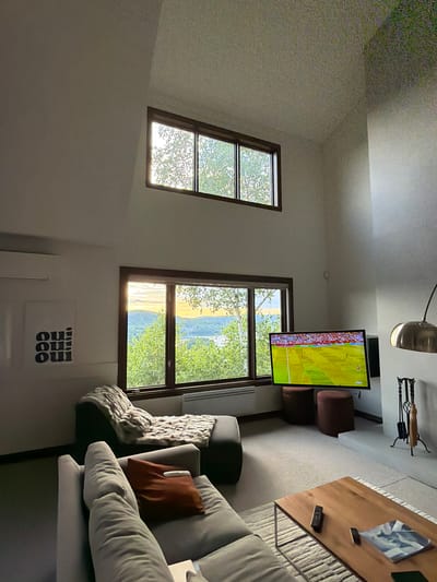 A living room in Mont-Tremblant, Canada, features a sectional sofa, a chaise lounge with a blanket, a large television displaying a soccer match, a coffee table with remotes and a magazine, and a fireplace set. Large windows offer a view of a body of water and trees, with the sun setting in the distance. A piece of art with the word "Oui" repeated is hung on the wall. The ceiling is high and angled, with a small window at the top. The room is well-lit by natural light and overhead lighting.