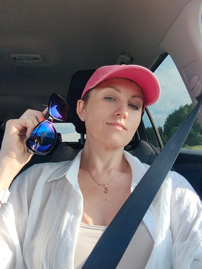 A woman is sitting in the driver's seat of a car, holding a pair of sunglasses in her left hand. She is wearing a pink baseball cap, a white open-collared shirt over a beige top, and a gold necklace with a small pendant. Her right hand is on the steering wheel, and a seatbelt is visible across her chest. Outside the car window, trees and a cloudy sky are visible. The image appears to be taken on a sunny day, possibly in a rural area given the presence of trees. The specific location within Юзуфоўскі сельскі Савет, Belarus, cannot be determined from the image alone.