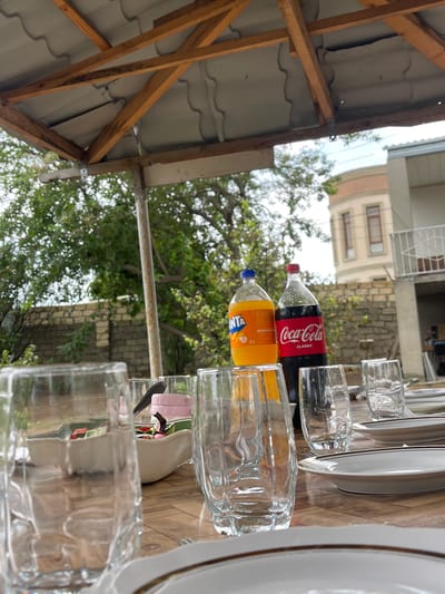 This image depicts a table set for a meal outdoors, likely in a backyard or patio area in Sabunchu Raion, Azerbaijan. The table is covered with a wooden surface and is set with several clear glasses, plates with brown rims, and a bowl containing a salad. Two large bottles of beverages, a Fanta and a Coca-Cola, are prominently displayed on the table. In the background, a brick wall and lush green trees are visible. A building with windows and a balcony can be seen further in the distance. The scene is captured from a low angle, looking upwards towards a wooden-framed roof structure. The lighting suggests it is daytime. There are no people visible in the image.
