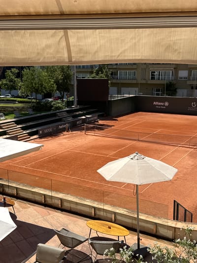 The image shows an outdoor clay tennis court in Palma, Spain. A shaded patio area with tables and chairs is in the foreground, with an umbrella providing shade over one of the tables. In the background, spectators' seating tiers are visible, with banners displaying sponsors such as "Allianz," "Oliver Raab," "ARXIE," and "Helvetia." Beyond the court, buildings with multiple windows and balconies are visible, surrounded by green trees. The sky is bright and clear, indicating a sunny day.
