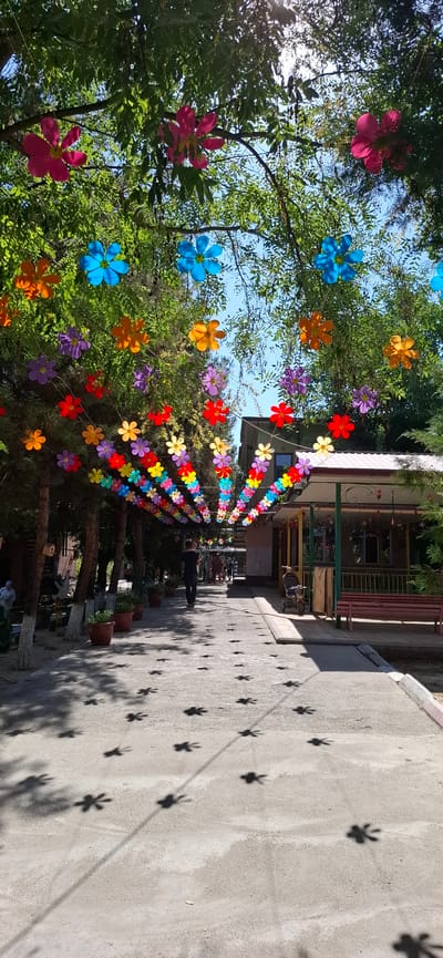 The image depicts an outdoor pathway decorated with strings of colorful paper flowers suspended between trees, creating a festive canopy. The pathway is paved and lined with potted plants on one side, and a building with a covered porch on the other. Several people are visible walking along the path, and a stroller is parked near the building. The ground displays numerous flower-shaped shadows, cast by the decorations above. The setting appears to be a public space in Sabzavot, Uzbekistan, possibly a park or a pedestrian area with shops or cafes. The lighting suggests it is daytime, with sunlight filtering through the leaves and decorations.
