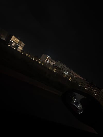 The image captures a nighttime scene, viewed from inside a vehicle, possibly from a passenger's seat looking out the side window. The perspective is at a steep diagonal angle, with the horizon line slanting upwards from left to right.

In the mid-ground, a series of multi-story buildings line what appears to be a street or pathway. Many windows in these buildings are illuminated with warm light, suggesting interior activity. The architecture appears modern, with visible balconies or external structures on some buildings. Further down the line of buildings, on the left side of the frame, one structure prominently displays visible text. The text clearly reads "LA VID RESTAURANT", indicating a commercial establishment.

The foreground is dominated by a dark strip, likely a road or ground, with faint vertical elements that could be small trees or decorative lighting poles extending along its edge. On the far right side of the image, a large, dark, and reflective surface is visible, which is unmistakably a car's side mirror. This mirror reflects a distorted view of the external environment, including some bright lights and blurred shapes, further confirming the perspective from within a vehicle.

The overall scene is very dark, indicative of a clear night. There are no visible people or specific activities other than the implied presence within the lit buildings. The setting appears to be an urban or suburban area in San Francisco Ocotlán (Ocotlán), Mexico.